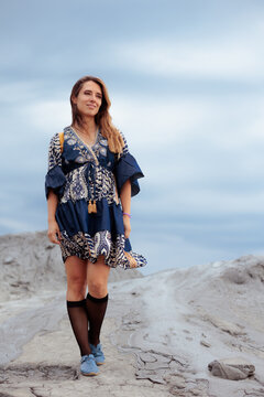 Attractive Young Woman Wearing Patterned Dress Smiling On Rocky Ground. Cheerful traveler posing outside looks forward stands on textured stone landscape confidently