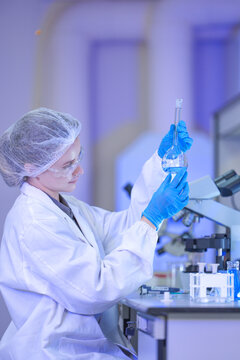 A female science professional in a white coat, protective eyewear and a sterile hairnet holding the flask level with her safety glasses at a well-equipped science laboratory bench.