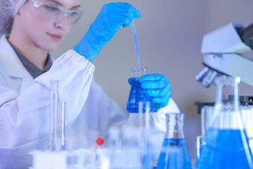 Fototapeta premium A woman researcher in white laboratory clothing, safety glasses and blue gloves holding the flask out beside the microscope at a brightly lit laboratory workbench.