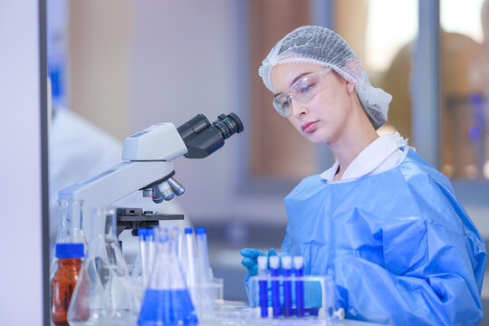 A woman laboratory scientist in a white lab coat, hairnet and clear safety goggles gripping the tube rack tight in both hands in a modern medical research laboratory.