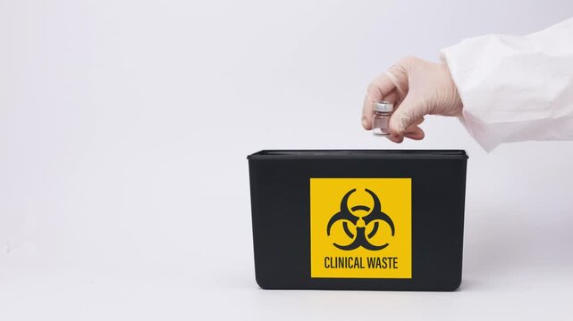Gloved hand carefully drops contaminated medical vial into black biohazard container for clinical waste hazard management and safe equipment disposal
