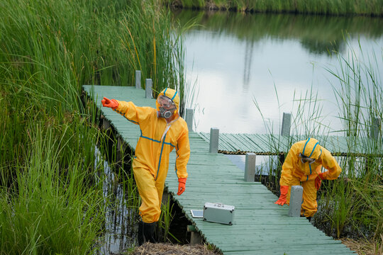 Hazmat suit respirator protective gear environmental inspection water sampling wetland marsh dock, two technicians collect sample near shoreline, showing contamination response and safety