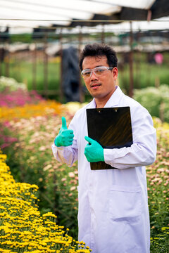 Researcher man giving thumbs up while holding clipboard in a flower nursery, wearing safety glasses, gloves and lab coat with confident positive expression
