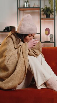 Sick young woman wrapped in plaid sitting, shivering from cold on sofa drinking hot tea in unheated apartment without heating due debt. Unhealthy lady in hat feeling discomfort try to warm up at home