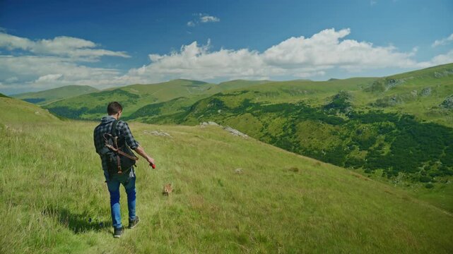 Man walks across grassy hill in the mountains with a rifle on his back and a dog nearby on a sunny day