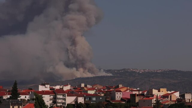 Massive wildfire sends dense smoke column above Vila Nova de Foz C&ocirc;a&rsquo;s rooftops, set against rugged hills under clear daylight. Dramatic static wide shot, warm sunlit atmosphere.