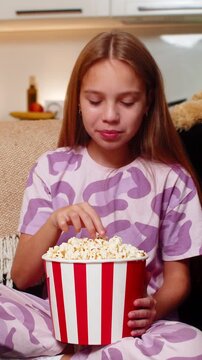 Teenager girl at night home watches movie takes popcorn from bowl laughs loud fun comedy scene. Caucasian child kid on sofa at evening chews snack smiles wipes tears of laugh enjoys leisure time joy