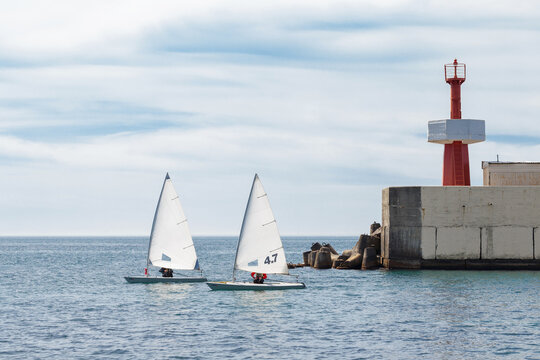 Two small dinghy sailboats with white sails race near stone breakwater and red lighthouse under cloudy sky