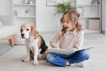 Fototapeta premium Cute little girl with Beagle dog reading book on floor in living room