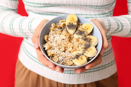 Young woman holding bowl of healthy oatmeal with banana on red background, closeup