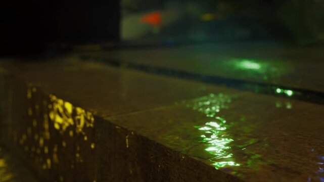 Water moves across polished stone, creating small ripples and reflections in a low light setting. The surrounding environment appears dark with hints of color from light.