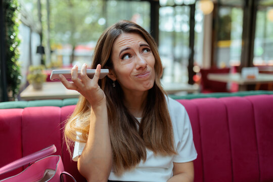 Confused Young Woman Listening To Voice Message On Her Smartphone. Doubtful thinking female model holding mobile phone near ear inside cozy bright restaurant