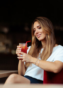 Smiling Woman Holding Refreshing Cocktail Indoors At the Bar. Happy charming lady sitting inside restaurant while enjoying cold delicious red beverage
