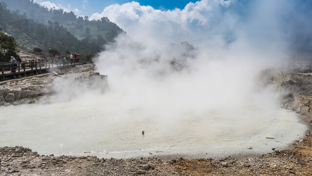 An acidic lake in the volcano&rsquo;s crater. Steam, evaporation rising from the water. Tourists walking along the wooden terrace. Green vegetation on the hillside. Blue sky. Indonesia. Java. Kawah Sikidang