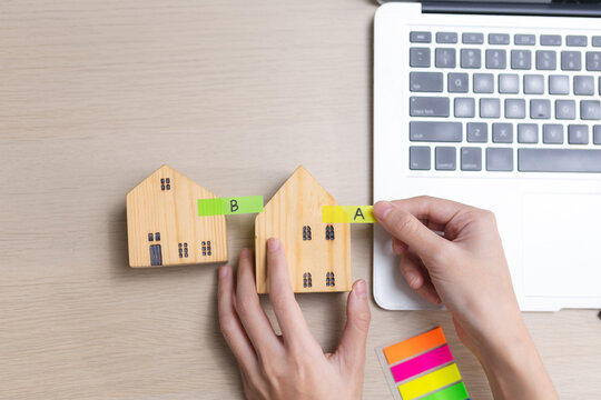 Property comparison and home buying concept. Person compares wooden house models labeled A and B beside laptop while evaluating real estate options, prices, housing decisions.