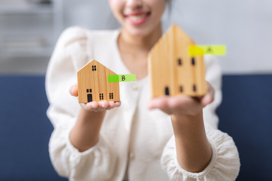 Property comparison, home buying concept. Person compares wooden house models labeled A and B while using laptop and notes to evaluate real estate options and make housing decision.