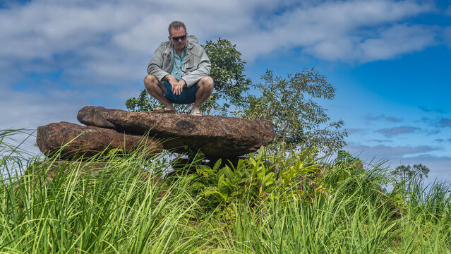 Large flat boulders are stacked in the shape of a pyramid. A man is crouching on the rocks, looking down. Lush green grass on the hill. Blue sky, clouds. Mauritius. La Vall&eacute;e Des Couleurs Nature Park 