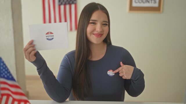 Woman smiling holds ballot envelope and points finger to voted sticker beside american flag at polling station; civic pride.