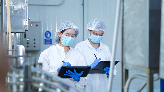 Female quality control inspectors writing notes on tablet and clipboard in food factory plant, Industrial production line operators checking quality control data in manufacturing facility