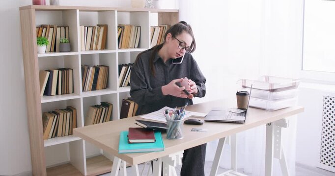 Corporate routine. Office makeup. Female manager talking on smartphone applying face powder at desk with laptop for business paperwork indoor.