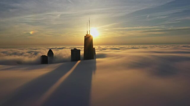 Cinematic aerial footage moving laterally across Chicago as the 875 North Michigan Avenue tower and surrounding skyscrapers emerge from a spectacular blanket of fog illuminated by the sun.