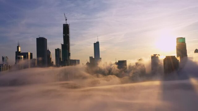 Sweeping aerial footage captures downtown Chicago skyscrapers emerging through rolling evening fog while brilliant sunset light creates a warm golden atmosphere across the city.