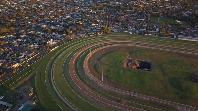 Newcastle Racecourse oval tracks and grassy infield beside dense residential suburbs, aerial medium orbit with soft golden glow contrast