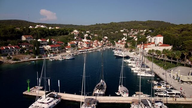 Maslinica, Croatia marina filled with sailboats beside red roof village, aerial ascend backdrop establishing overview of promenade as people walk along coast