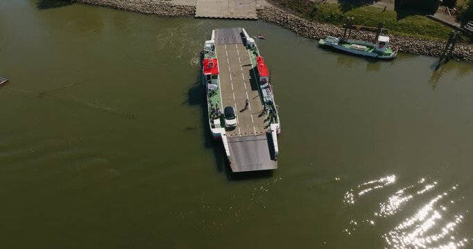 Ferry departs from a riverside dock with a car passenger aboard, crossing the Dutch Lek River through a typical flat polder landscape under a clear blue summer sky.