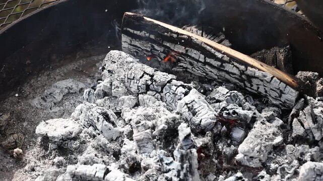 Close-up of charred firewood and glowing embers covered in white ash, with faint smoke rising from the remains of a wood fire.