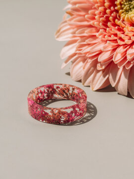 Handmade epoxy resin ring with pink dried flowers beside blossom on neutral background.