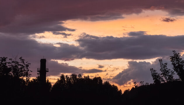 Dramatic orange sunset behind dark clouds and tree silhouettes, atmospheric urban landscape.