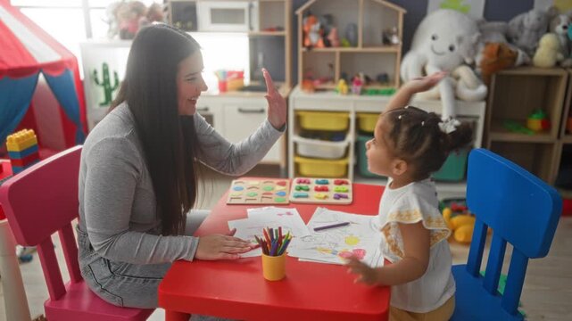 Woman raises hand for high five with little girl over crayons and coloring sheets at preschool building; playful learning.