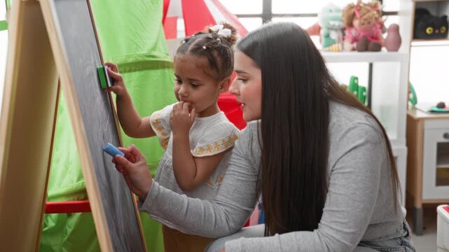 Woman teacher drawing with blue chalk on easel while little girl holds eraser and fingers at mouth in preschool; curiosity learning.