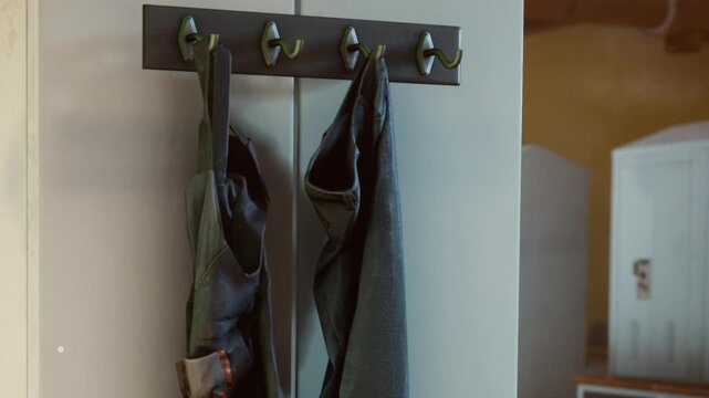 Two items of clothing are hanging on hooks placed on a wall in a room. The setting is casual and indoor. The colors of the clothing are dark and simple.