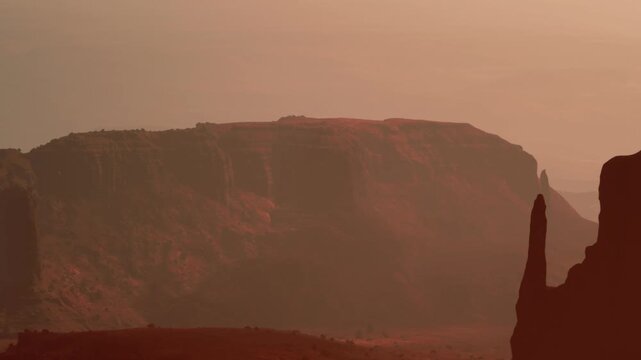 A warm sunset creates shadows on a rocky landscape. The view shows cliffs and distant mountains surrounded by flat desert land. The colors change as day shifts to night.