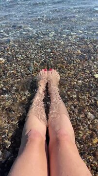 Women's legs relaxing in clear shallow sea water over colorful pebbles on a sunny beach