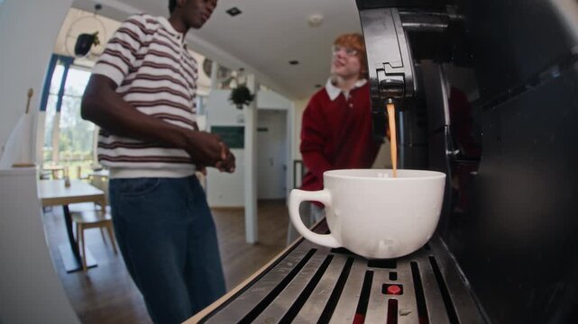 Coffee machine pouring espresso into cup in foreground while African American and Caucasian young adult male coworkers chat in background. Ideal for corporate culture and office breakroom videos