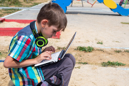 A cute boy schoolboy dressed in casual clothes is engaged on a laptop on the playground, a schoolbag lies nearby