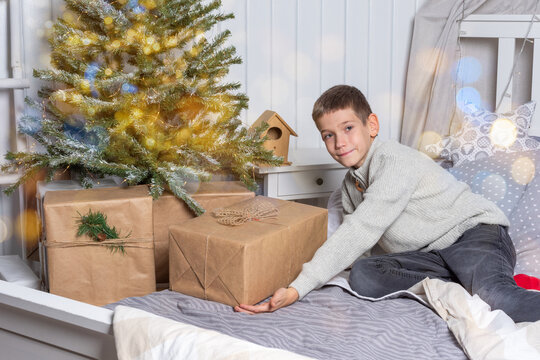 Cute teenage boy in a sweater sitting on the bed and putting presents under the Christmas tree. New Year's Eve