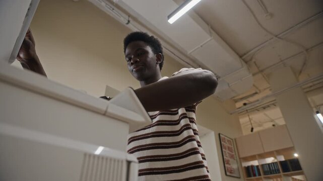 Young adult African American man using office copier machine, scanning documents, and collecting printed papers from low angle. Footage is suitable for corporate training or workplace tech ads