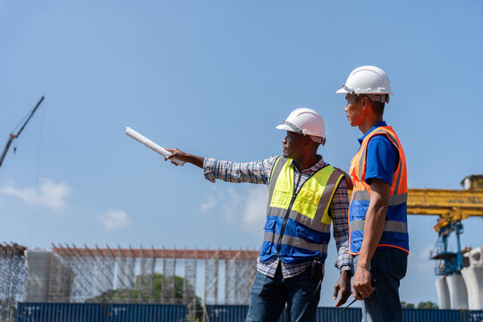 Construction engineers team with blueprint pointing at building site outdoors, Civil engineers discussing project plan at site, Construction workers inspecting development site