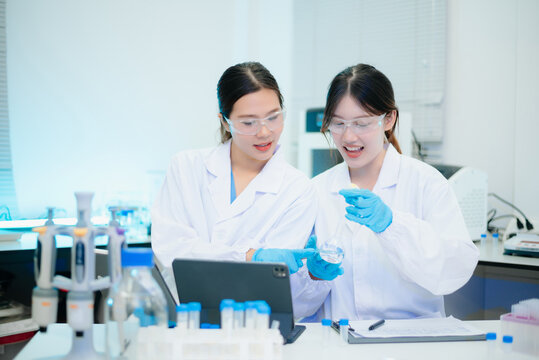 Two smiling Asian female scientists analyzing chemical liquid in a flask while using a digital tablet in a bright biotechnology laboratory.