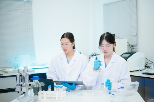Two smiling Asian female scientists analyzing chemical liquid in a flask while using a digital tablet in a bright biotechnology laboratory.