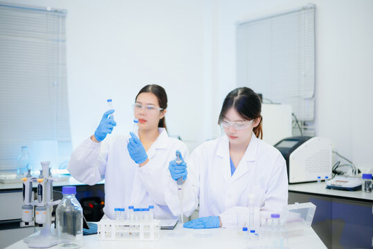 Two smiling Asian female scientists analyzing chemical liquid in a flask while using a digital tablet in a bright biotechnology laboratory.