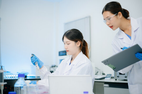 Two Asian female scientists collaborating with a micropipette and laptop computer during research in biotechnology laboratory.