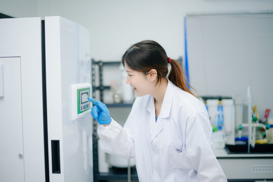 Asian female scientist setting parameters on a digital control panel of a storage unit in a modern biotechnology laboratory.