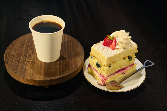 Piece of fruit cake on a plate with a spoon and black coffee in a disposable cup on a brown stand