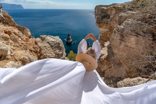 Woman hat dress, woman wearing summer hat and flowing white dress stands on a rocky cliff overlooking the ocean