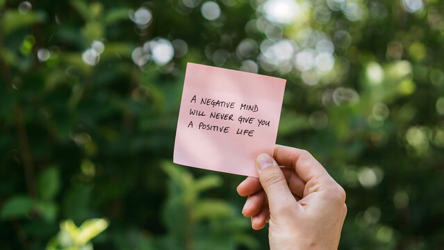 A hand holding a pink sticky note with positive life advice in the garden